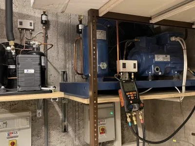 Zwei Verdichter auf einem Stahlgestell mit Kupferleitungen, davor ein digitales Manometer, darunter Schaltkästen mit KGS-Aufklebern