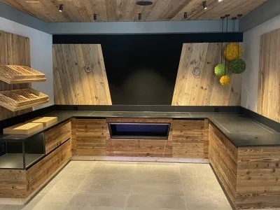 Theke aus Altholz mit dunkler Arbeitsplatte und eingelassener beleuchteter Kühlvitrine, links Körbe in einem Regal