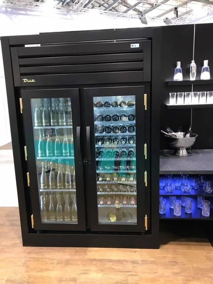 Schwarzer Kühlschrank mit zwei Glastüren, darin Flaschen und Wein, daneben ein Regal mit Gläsern
